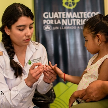Mujer joven con trenza toma muestra de la mano de una niña frente a un cartel azul con el texto 'Guatemaltecos por la Nutrición'