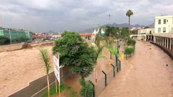 Río con agua marrón y turbia desbordado sobre un parque con senderos, árboles y un edificio claro, bajo un cielo nublado con cerros al fondo