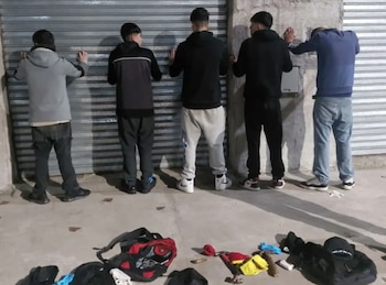 Cinco hombres jóvenes de espaldas, con las manos apoyadas en una persiana metálica gris y una pared de concreto. En el suelo hay mochilas y prendas de vestir