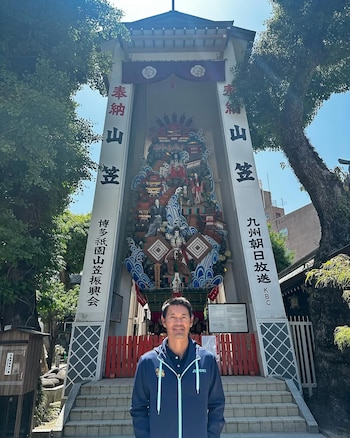 Un hombre con chaqueta azul claro y oscuro sonríe frente a una torre alta con decoración tradicional japonesa, textos en idioma japonés y árboles a los lados
