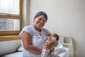 Una mujer sonriente, con blusa estampada, sostiene a un bebé en brazos. El rostro del bebé no es visible. Al fondo, una ventana y una pared clara