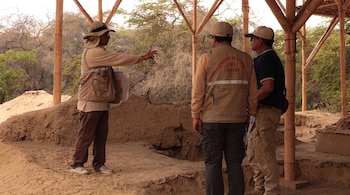 Tres personas con gorra y chaleco están junto a un terreno elevado de arena bajo postes de bambú en una zona arbolada.
