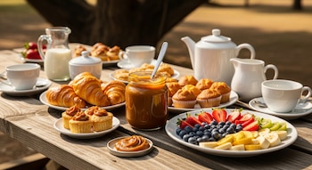 Mesa de madera con frasco de dulce de leche, leche, tazas blancas, croissants, magdalenas y un plato con fresas, arándanos, kiwi y plátano.