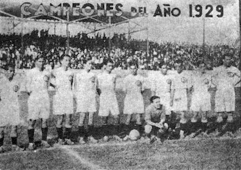 Fotografía en blanco y negro de un equipo de fútbol formado por diez hombres de pie y uno arrodillado en un campo, con una multitud al fondo