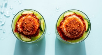 Toma cenital de dos chamoyadas de pepino en vasos, con hielo, chamoy, chile en polvo y rodajas de pepino. Fondo azul claro con gotas de agua.