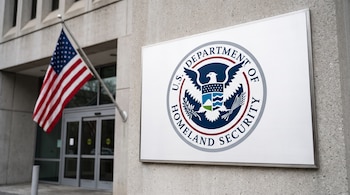 Placa con escudo del Departamento de Seguridad Nacional de Estados Unidos y su águila calva, bandera estadounidense, edificio de concreto y puertas de vidrio.