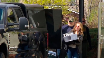 Una mujer con gafas de sol sale de un edificio llevando un equipo electrónico, con un hombre barbudo detrás y una camioneta oscura estacionada a un lado