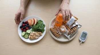 Manos de adulto sobre mesa de madera clara eligiendo entre plato saludable (frutas, verduras, pollo) y otro con comida ultraprocesada. Glucómetro visible.