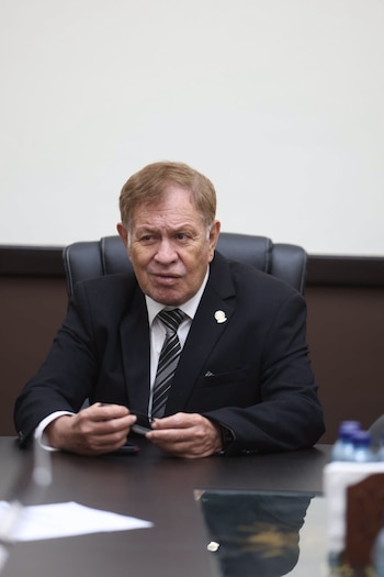 Hombre de edad con traje oscuro, camisa blanca, corbata a rayas y cabello castaño rojizo, sentado en una silla negra frente a una mesa oscura. Sostiene un bolígrafo