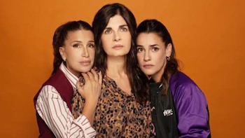Tres mujeres posan de frente con expresiones serias sobre un fondo naranja. La mujer izquierda tiene coleta y chaleco; la del centro, pelo suelto y blusa estampada; la derecha, coleta y chaqueta