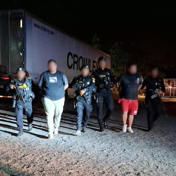 Varias personas con rostros borrosos permanecen sentadas a una mesa mientras agentes de policía las supervisan de noche