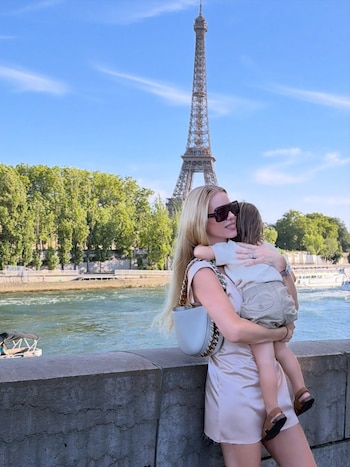 Una mujer rubia con gafas de sol y vestido claro sostiene a un niño. Están sobre un muro junto a un río, con la Torre Eiffel y árboles al fondo