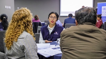 Mujer con gafas y chaleco de ONPE en mesa con papeles, hablando con otras dos personas sentadas de espaldas. Al fondo, una pantalla de proyección y un logo de PNUD