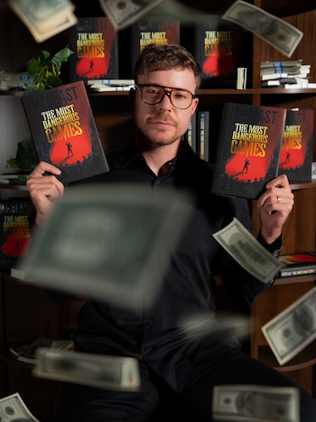 Un hombre con gafas y camisa negra sostiene dos libros con cubierta roja mientras billetes de dólar flotan en el aire