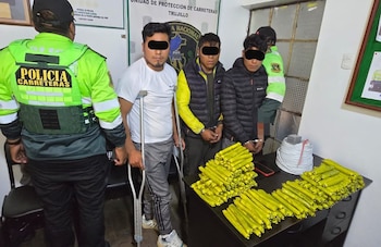 Tres hombres bajo custodia policial permanecen de pie junto a una mesa con decenas de cartuchos amarillos y un rollo de cable