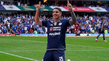 Un futbolista con uniforme azul y el número 21 celebra con las manos levantadas y los dedos cruzados en un estadio lleno de espectadores.