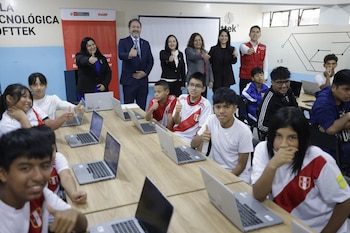Sala con diez estudiantes y siete adultos haciendo pulgar arriba. Computadoras portátiles en escritorios de madera. Pantalla de proyector y logos en la pared