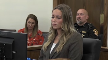 Una mujer joven rubia vestida con saco oscuro permanece sentada frente a un monitor en un tribunal, con un oficial al fondo