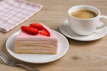 Rebanada de pastel de capas rosa y trozos de fresa en un plato blanco junto a un tenedor y una taza de café sobre una mesa de madera.