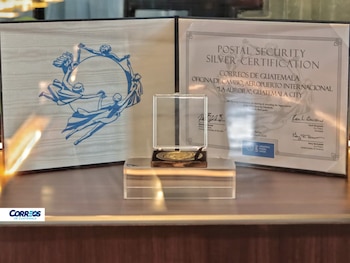 Una Certificación de Seguridad Postal de Plata enmarcada junto a una placa con figuras circulares y un medallón en una vitrina transparente, sobre una superficie de madera