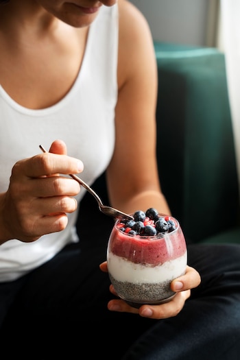 Persona con camiseta blanca que sostiene un vaso transparente con capas de semillas de chía, yogur, licuado rojo y arándanos