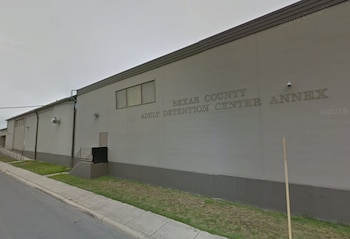 Fachada de un edificio gris con el texto Bexar County Adult Detention Center Annex grabado en el muro, junto a una acera y una calle