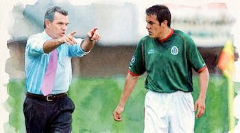 Dos hombres en un campo verde. Uno de camisa azul claro y corbata rosa señala con el dedo. El otro viste camiseta de fútbol verde y pantalón blanco.