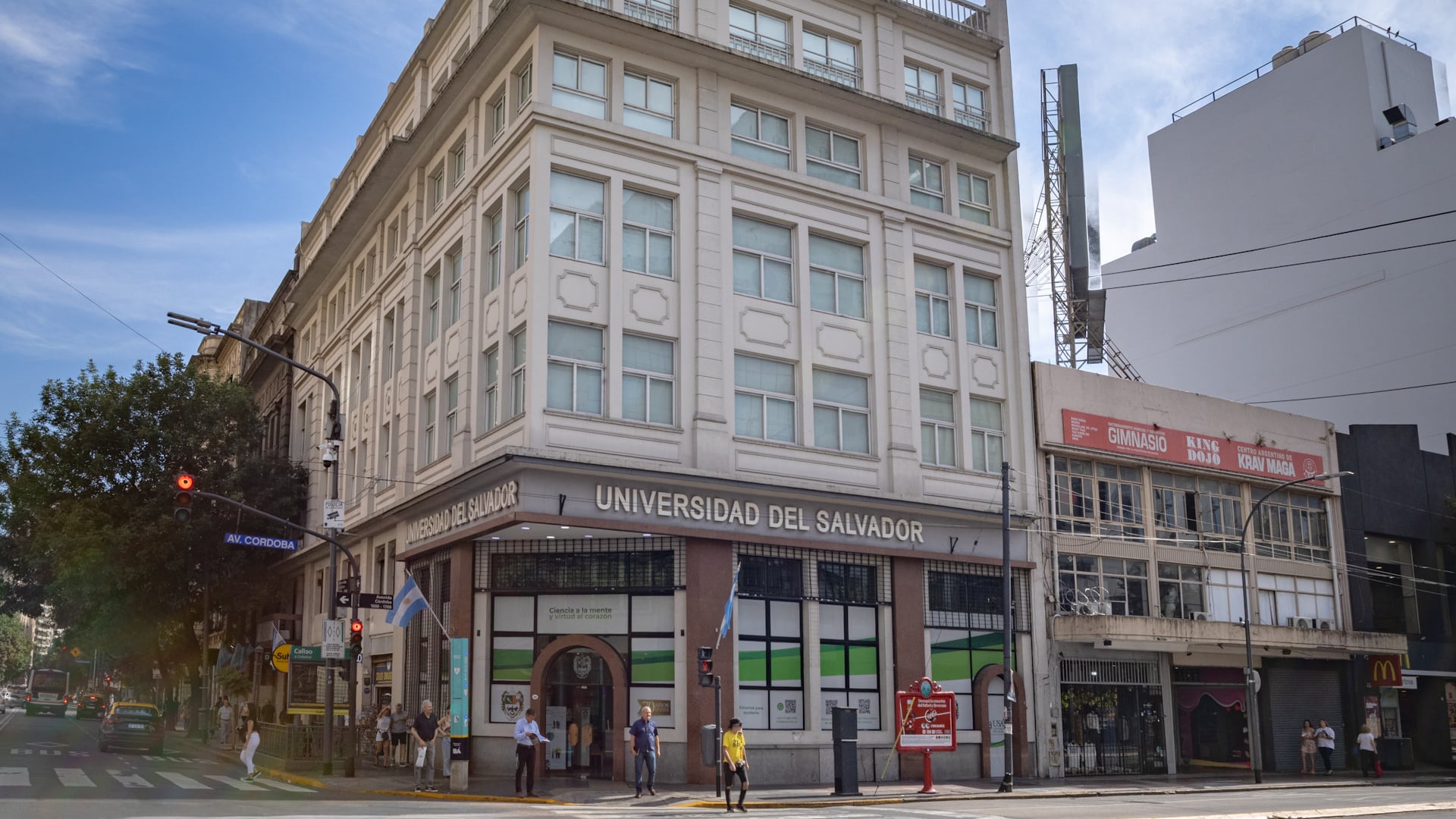 Peatones y vehículos transitan por la Avenida Córdoba en Buenos Aires, frente a la imponente fachada de la Universidad del Salvador en un día soleado.