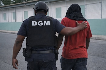 Vista trasera de un hombre con uniforme DGM y casco, su brazo sobre el hombro de un hombre con camiseta roja y cabeza cubierta, en una calle.