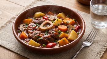 Plato hondo con guiso de osobuco, papas, zapallo, zanahorias, arvejas, pimientos y hongos. Tenedor y vaso de agua al lado, sobre individual y mesa de madera.