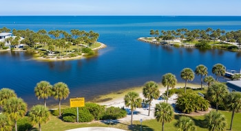 Vista aérea de Ballard Park, Florida: palmeras, césped verde, agua azul oscuro, pequeñas playas de arena y un letrero amarillo de 'Warning' bajo cielo azul claro.