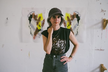 Una mujer mira de frente, viste gorra oscura, camiseta negra con tigre, jeans, collar plateado y reloj. Detrás hay una pared blanca con dos cuadros abstractos