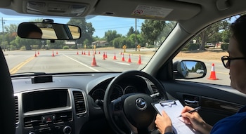 Vista interior de un coche durante un examen de conducir, mostrando a un examinador con un portapapeles y una carretera con conos de tráfico.