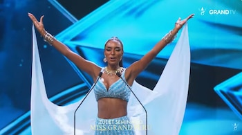Una mujer con traje azul y velos blancos alza los brazos frente a dos micrófonos en un escenario iluminado de azul