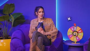 Ashley Grace sentada en un sillón morado sostiene un micrófono con ambas manos, junto a una planta y una ruleta multicolor