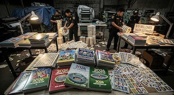 Agentes de policía inspeccionan una imprenta clandestina con montones de álbumes y láminas de figuritas falsas del Mundial 2026. Hay un cartel que indica 'S/9 MILLONES VALOR ESTIMADO'.