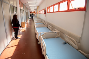 Pasillo de hospital con hilera de camas médicas blancas y colchones azules a la derecha. Una mujer de espaldas está a la izquierda; personal se ve al fondo