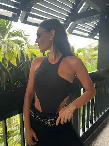 Mujer de cabello oscuro y gafas de sol, con body negro de recortes y cinturón de tachuelas, posa de perfil en un balcón con vegetación tropical