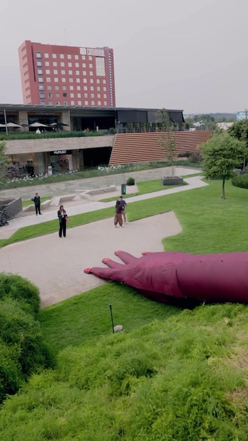 Mano inflable de color rojo oscuro sobre el césped. Sendero con personas caminando. Edificios de arquitectura moderna y un hotel Camino Real rojo en el fondo