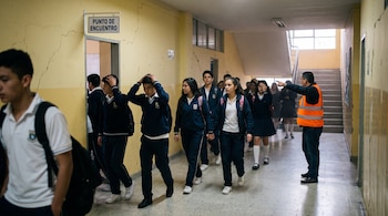 Estudiantes con uniforme caminan por un pasillo con grietas mientras un hombre con chaleco reflectante señala la salida.
