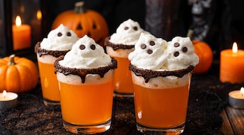 Cuatro vasos con bebida naranja, crema batida decorada como fantasmas con chispas de chocolate, bordes de chocolate, calabazas y velas encendidas.