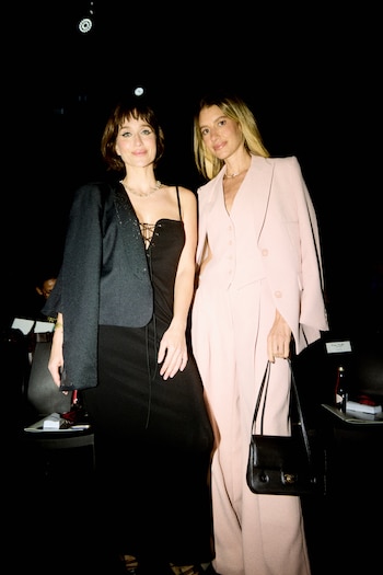 Dos mujeres posan de pie; una viste un vestido negro con saco oscuro y la otra lleva un traje de color rosa claro con una cartera negra