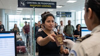 Una mujer frente a una ventanilla de migración extiende un pasaporte marrón de Ecuador hacia un oficial con guantes.
