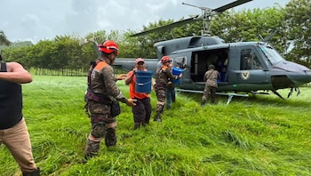 Un helicóptero gris en un campo de hierba alta. Seis personas, algunas con uniforme, descargan cubos azules del helicóptero. Al fondo, árboles y cielo nublado
