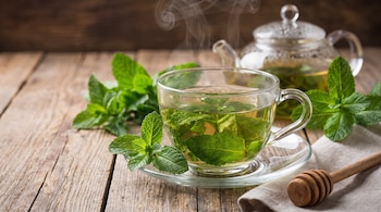 Una taza de té de menta humeante con hojas frescas, una tetera de vidrio, un dipper de madera y un paño de tela sobre una mesa de madera.