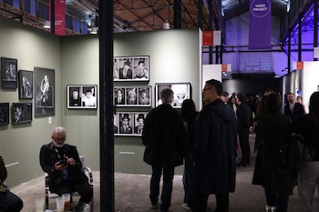 Personas de pie y sentadas frente a paneles con retratos fotográficos en blanco y negro dentro de una sala de exposiciones techada