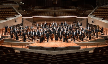Integrantes de una orquesta están de pie con sus instrumentos en el escenario frente a las butacas vacías de un auditorio