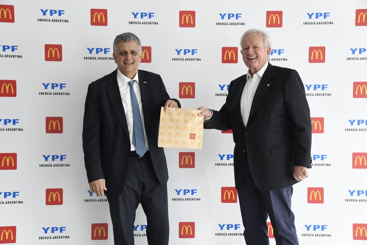 Un funcionario y un empresario intercambian una bolsa de McDonald's en un evento de las marcas YPF Energía Argentina y McDonald's, con sus logotipos repetidos de fondo.