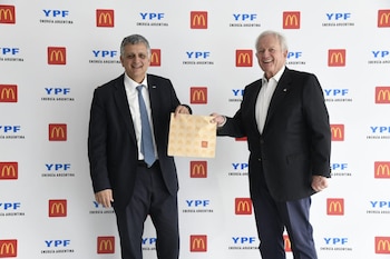 Dos hombres de traje se sonríen y extienden una bolsa de papel marrón con el logo de McDonald's, frente a una pared blanca con logotipos repetidos de YPF y McDonald's