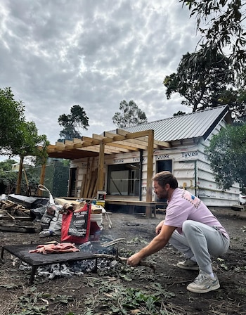 Hombre agachado junto a una parrilla con carne y carbón, en un patio trasero. Detrás, una casa en construcción cubierta con Tyvek y una pérgola de madera